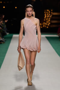 Model walking a runway in a pale pink one-shoulder sculpted mini dress, holding a beige clutch.