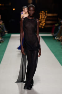 Model walking a fashion runway at night in a sheer black ensemble with draped scarf-like fabric flowing from the hip portion of the dress.