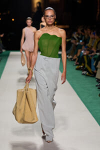 Model on a runway wearing a green strapless top and light gray wide-leg pants, carrying a large beige tote bag.
