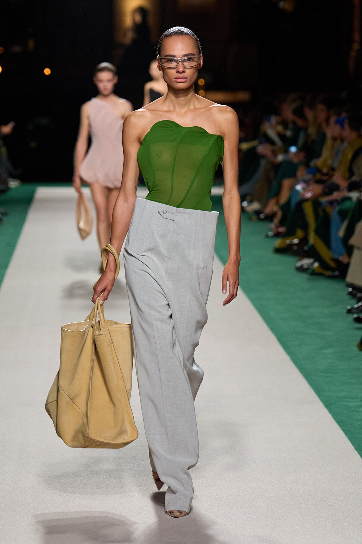 Model on a runway wearing a green strapless top and light gray wide-leg pants, carrying a large beige tote bag.