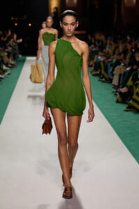 Model walks a fashion show wearing a green one-shoulder dress with a draped, bubble hem and brown loafers, carrying a brown clutch on the runway.