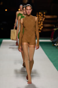 Model walks the runway in a tan-brown lace bodysuit with cold-shoulder design and beige thigh-high stockings.