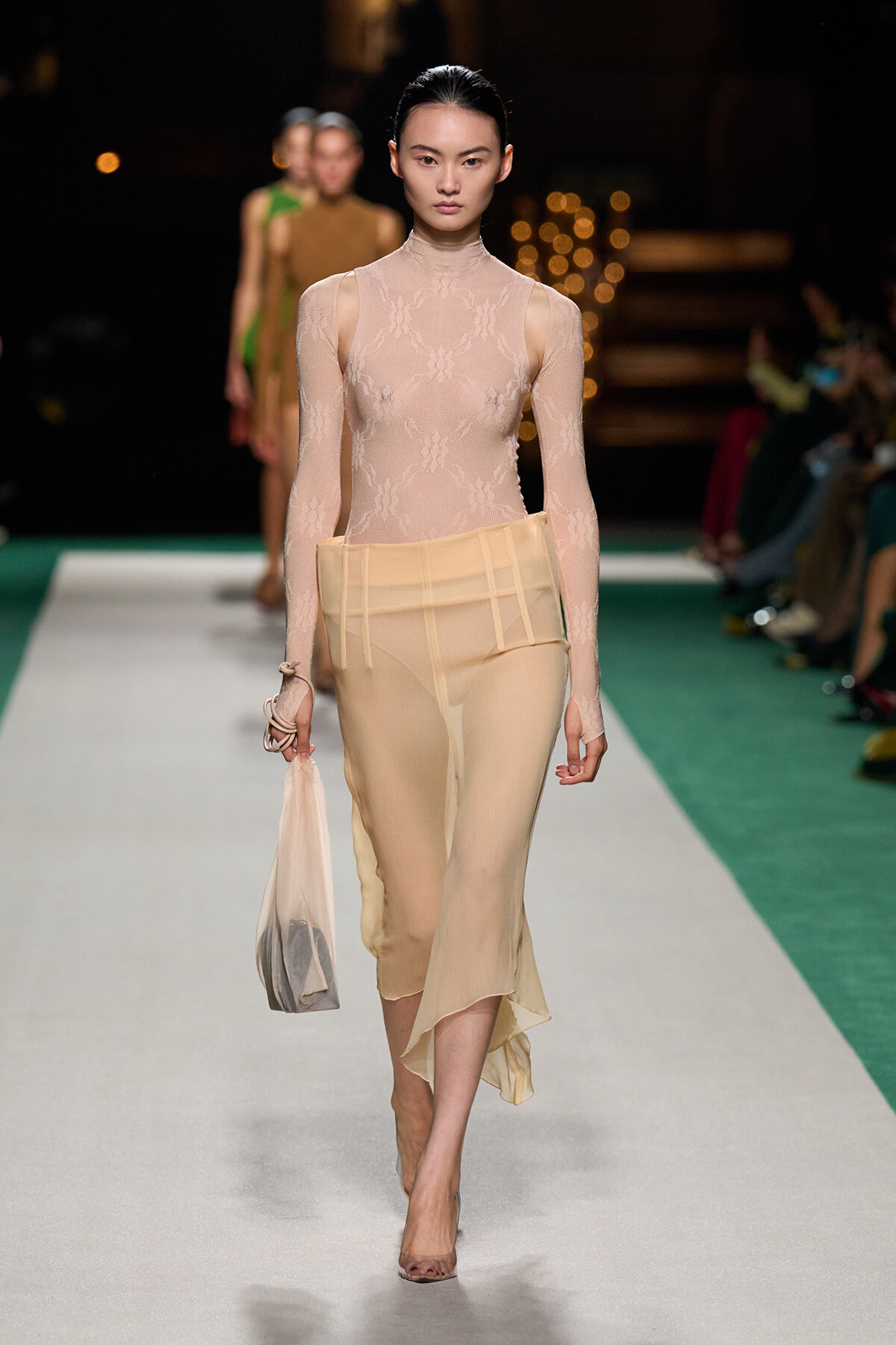 Model walks the runway in a nude lace turtleneck bodysuit and a pale sheer skirt, carrying a small beige bag.