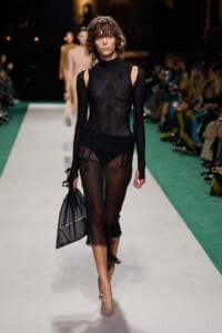 Model walking a fashion runway in a sheer black lace dress with cutout shoulders, holding a black mesh bag.