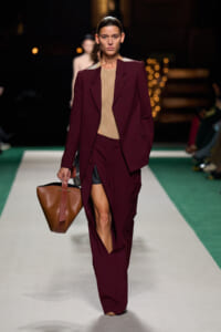 Model walks a fashion runway wearing a burgundy suit with an oversized blazer and wide-leg pants, beige lace top, and a brown leather tote bag.