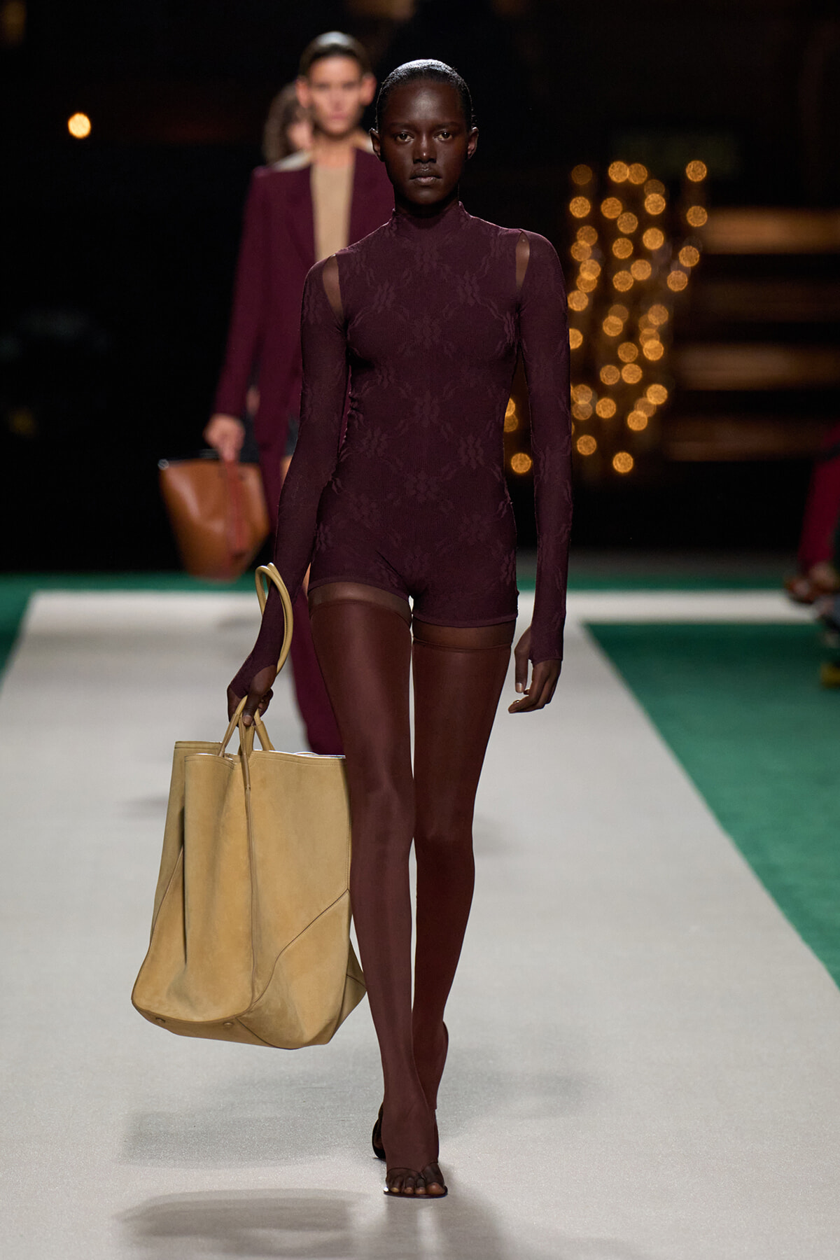 Model walks the runway in a burgundy lace long-sleeve bodysuit, carrying a large beige tote bag on her left side.
