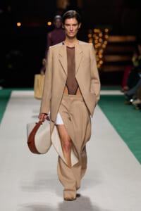 Model walks the runway in a beige oversized suit with a sheer brown bodysuit, carrying a round beige and brown bag.