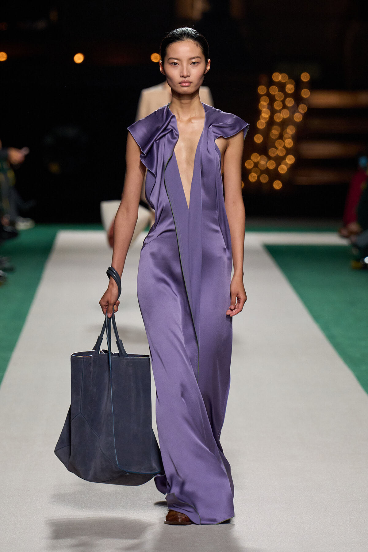 Model walks a purple satin jumpsuit with deep V neckline and ruffled shoulders on a runway, carrying a large dark tote bag.