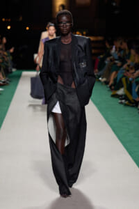 Model walks a runway in a black satin blazer, sheer top and oversized dark trousers with a high slit showing white underlayer.