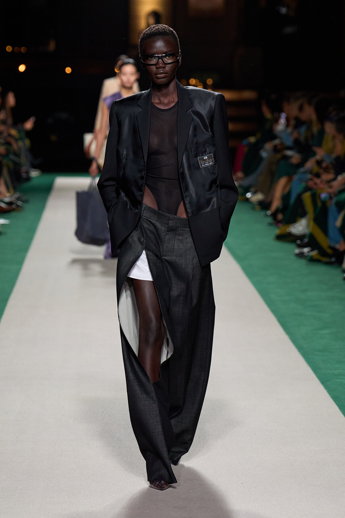 Model walks a runway in a black satin blazer, sheer top and oversized dark trousers with a high slit showing white underlayer.
