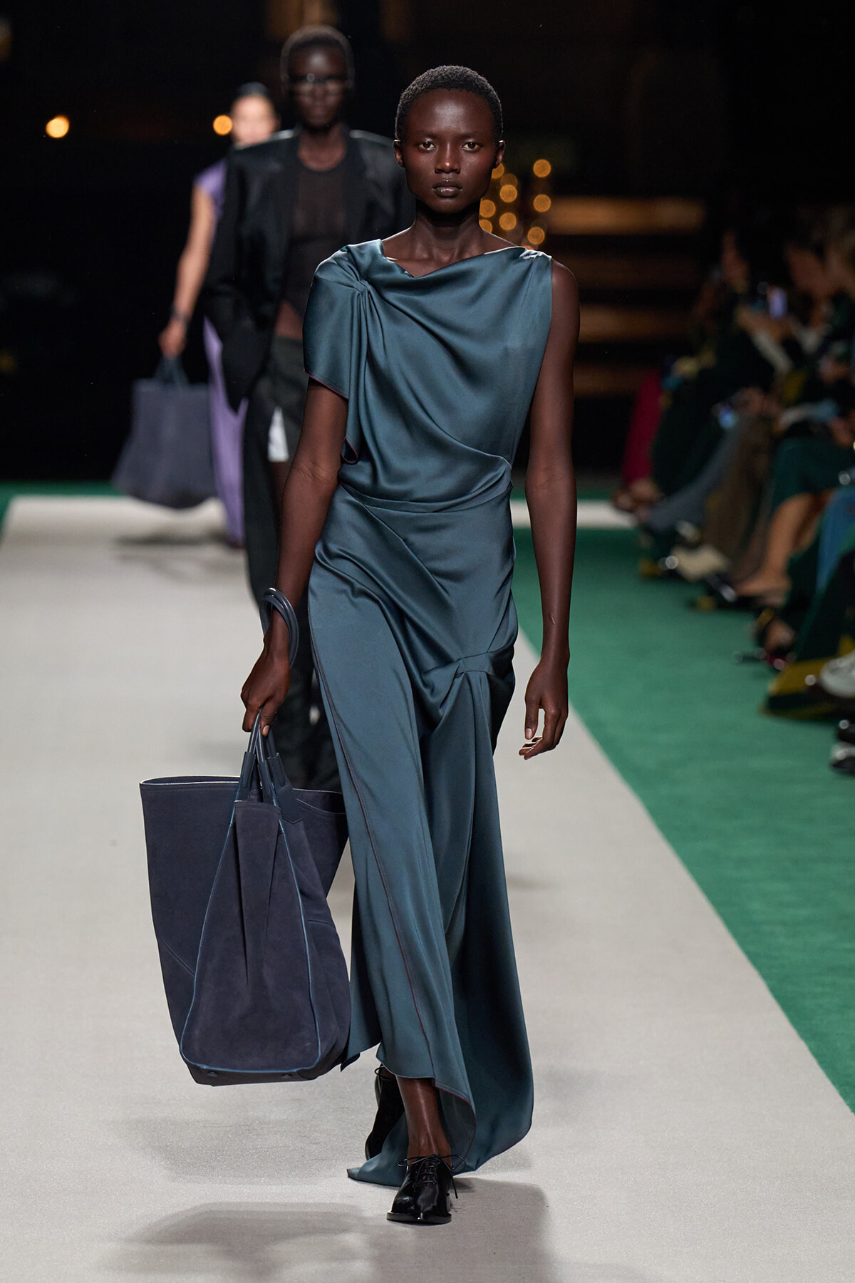 Model walking a fashion runway in teal satin draped jumpsuit, carrying a large navy tote bag.