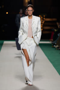Runway model in an oversized white suit with a split-front trouser, pale pink top, and rectangular glasses.
