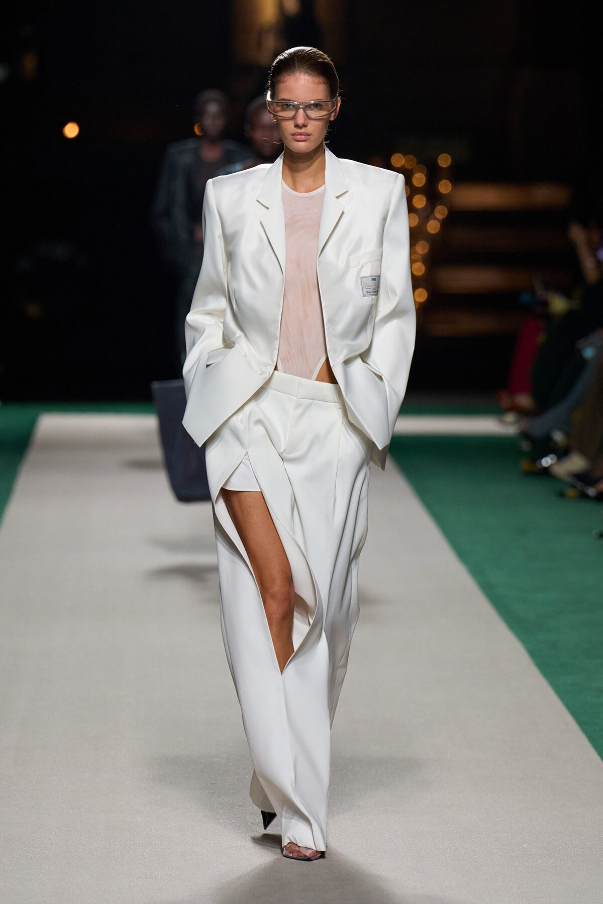 Runway model in an oversized white suit with a split-front trouser, pale pink top, and rectangular glasses.
