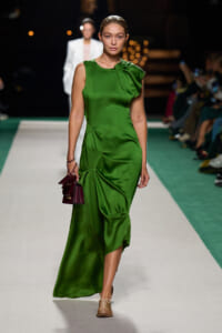 Model walking a runway in an emerald green satin dress with draped detail, carrying a small burgundy handbag.