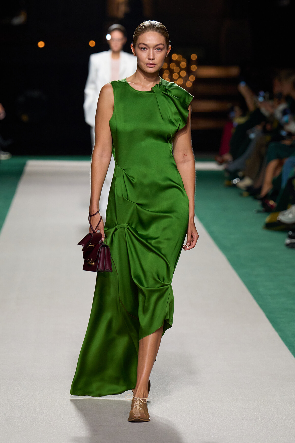 Model walking a runway in an emerald green satin dress with draped detail, carrying a small burgundy handbag.