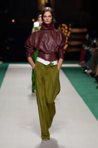 Model walking a runway in a burgundy leather high-neck top with voluminous sleeves, paired with olive green wide-leg trousers and a white belt.