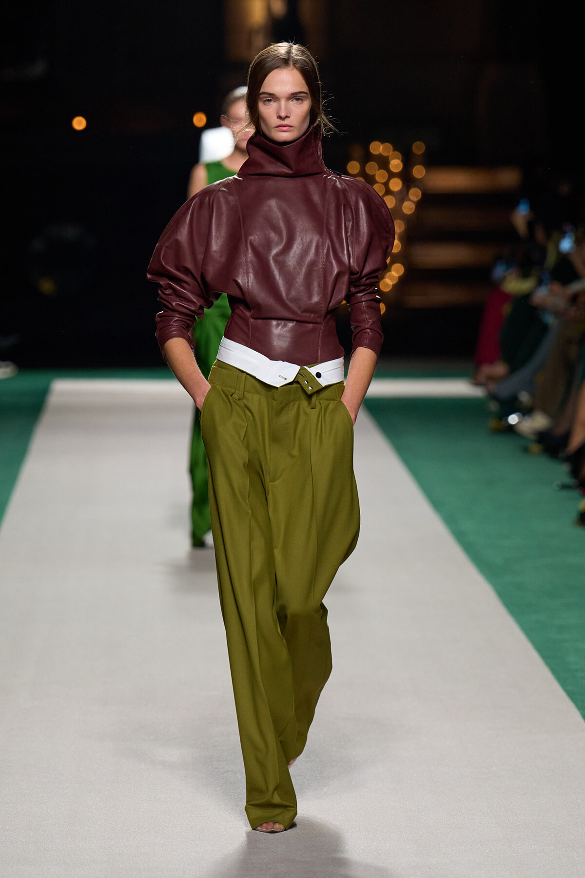 Model walking a runway in a burgundy leather high-neck top with voluminous sleeves, paired with olive green wide-leg trousers and a white belt.