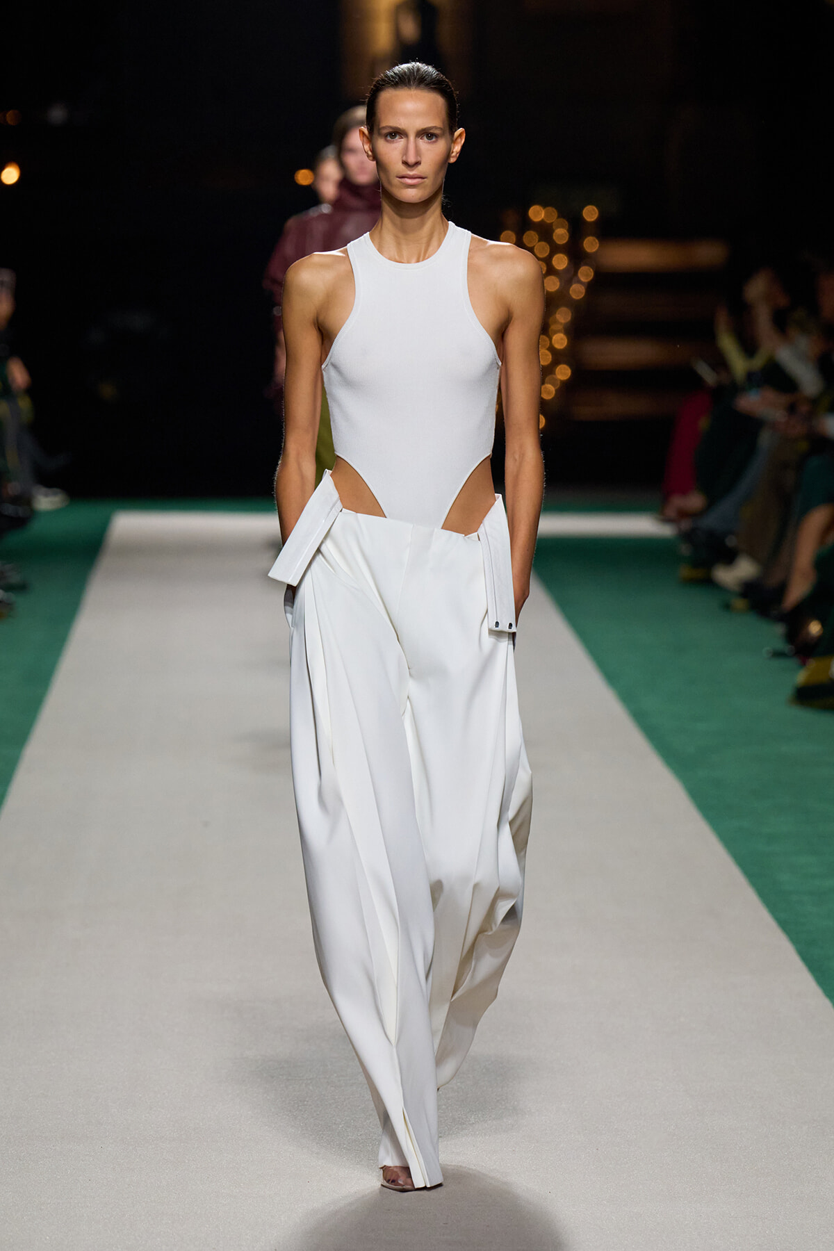 Model walking the runway in an all-white ensemble: sleeveless high-neck bodysuit with hip cutouts and wide, pleated white trousers with attached panels at the hips.
