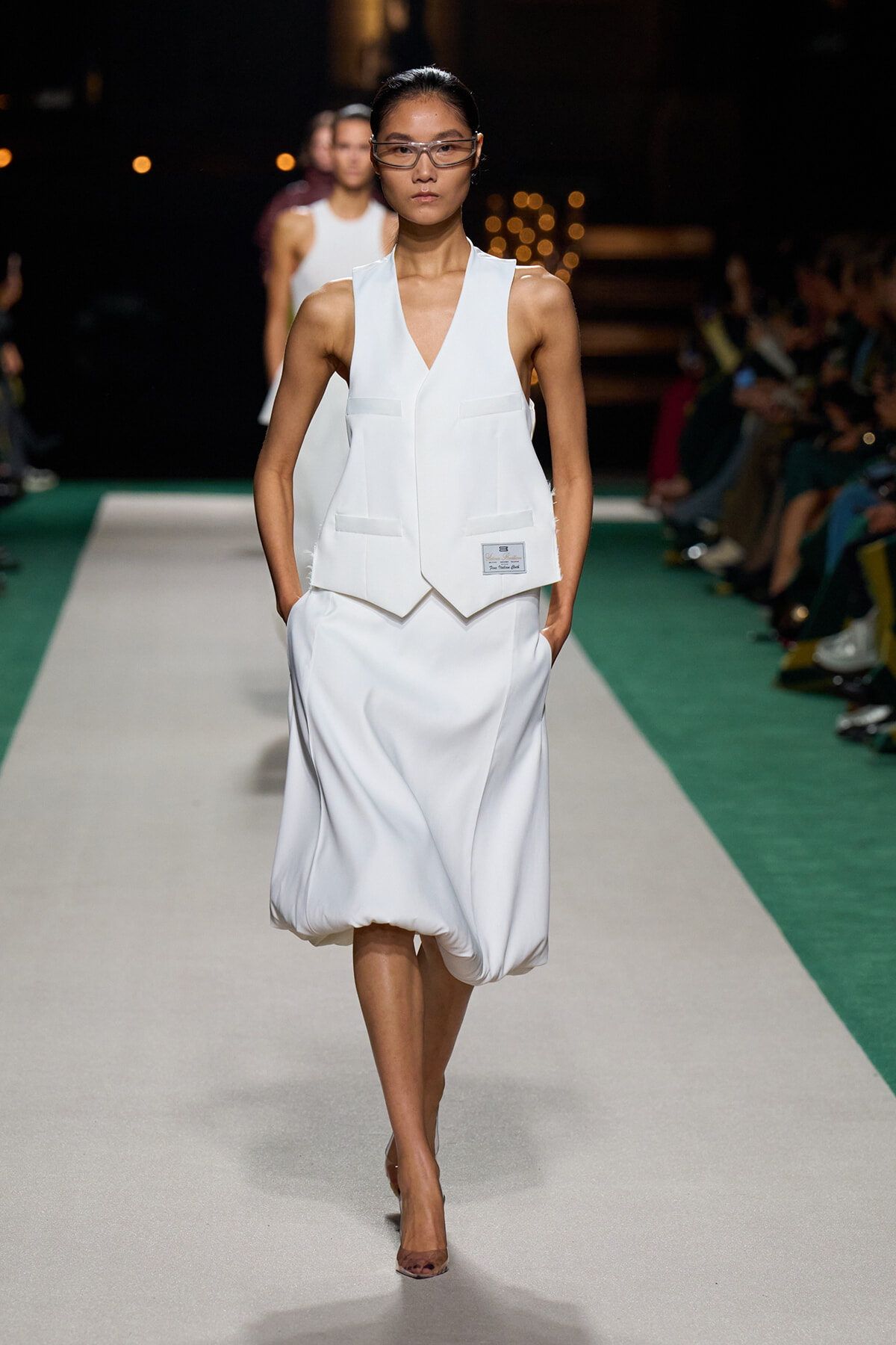 Model walks the runway in a white sleeveless vest over a bubble-hem dress, wearing clear protective eyewear.