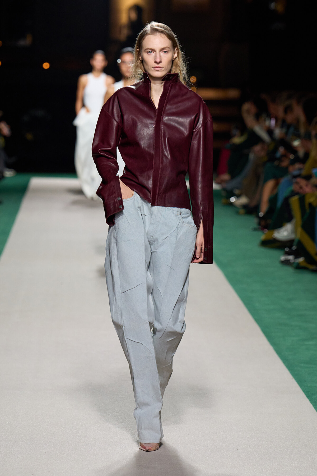 Model walks a fashion runway in a burgundy leather zip-up jacket paired with oversized light gray trousers; audience in the background blurred.