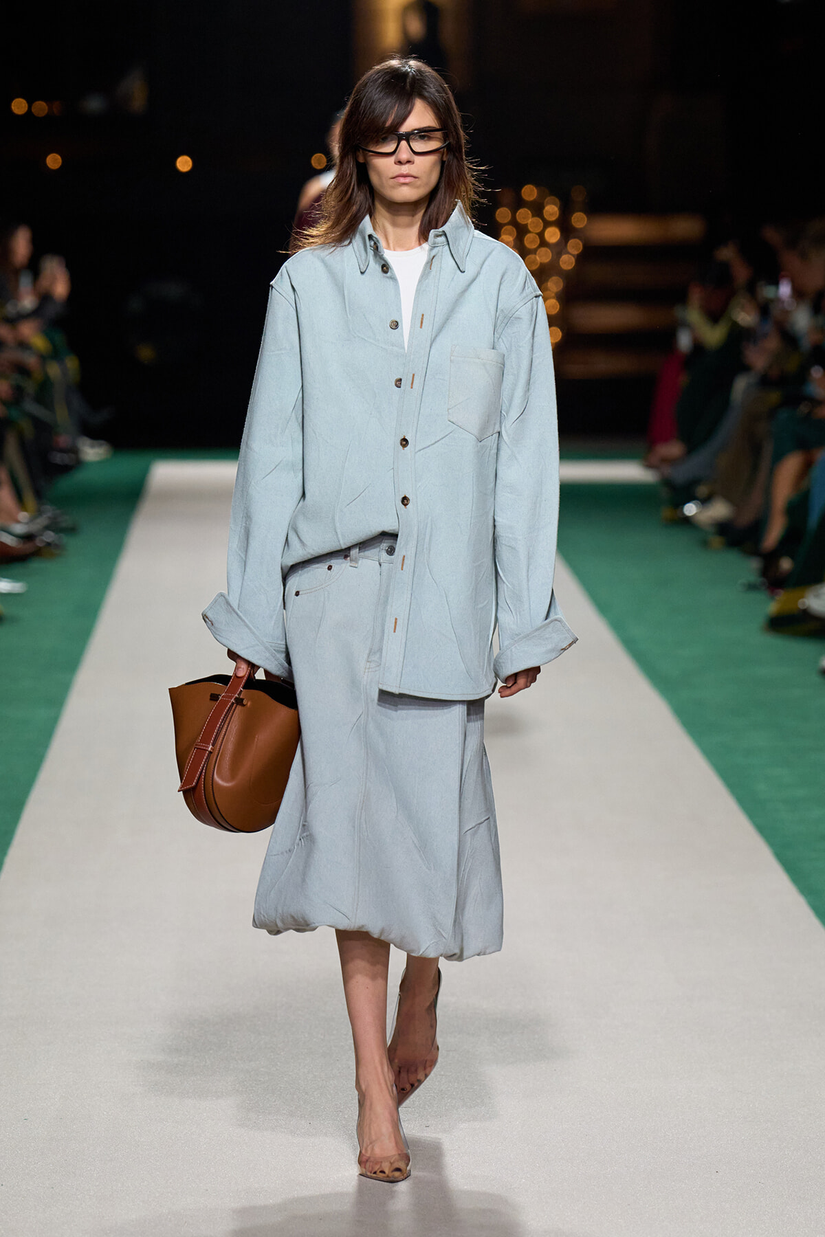 Female model walks the runway in an oversized light-blue denim shirt and matching midi skirt, carrying a brown leather handbag.