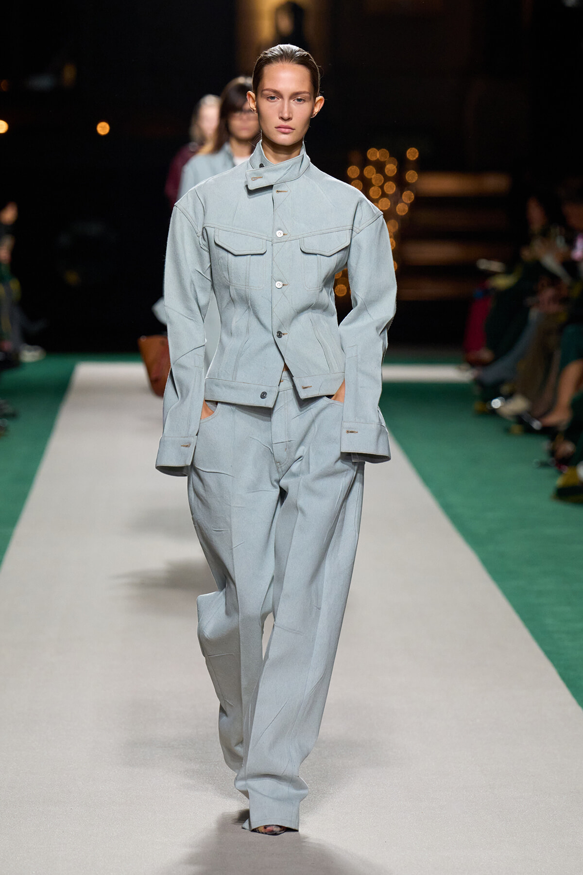Model walks the runway in a light blue tailored jacket and matching wide‑leg trousers with a utilitarian look on a fashion runway.