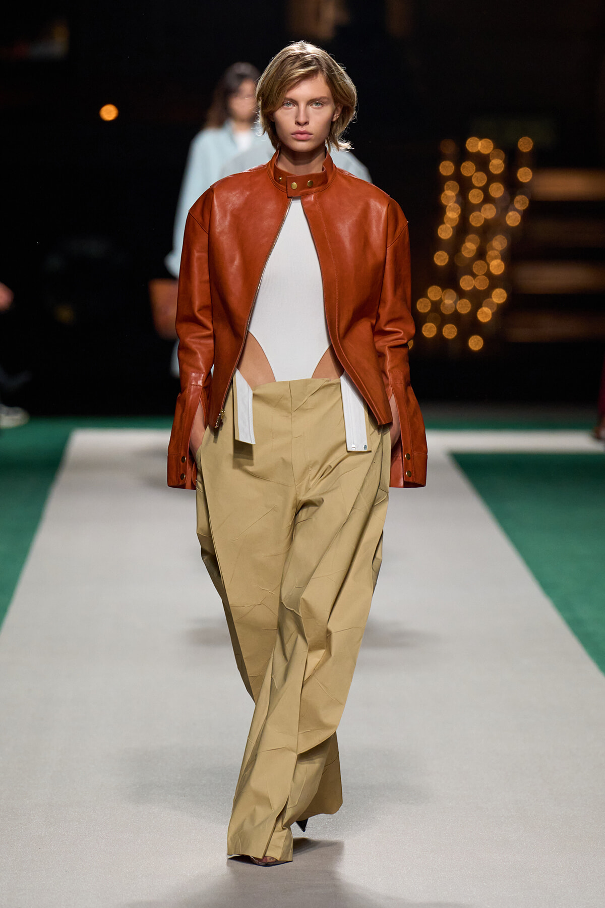 Model on a runway wearing a rust-colored leather jacket over a white bodysuit with oversized tan trousers on a fashion show runway.