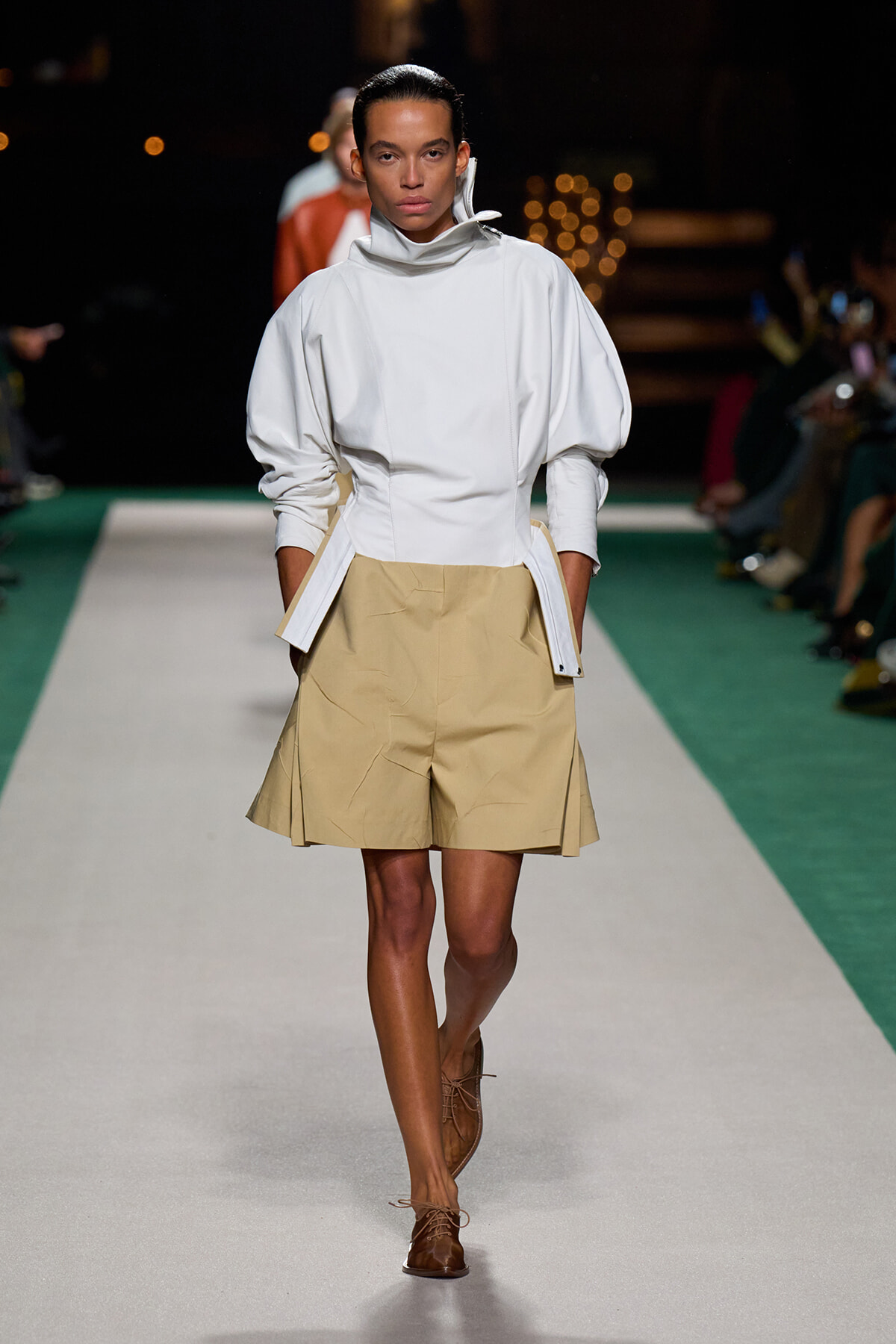 Model walking a runway in a white high-neck, puff-sleeve top and tan pleated shorts with brown lace-up shoes.