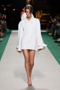 Model walking a fashion runway in an oversized white coat with wide cuffs and short hem, neutral backdrop.