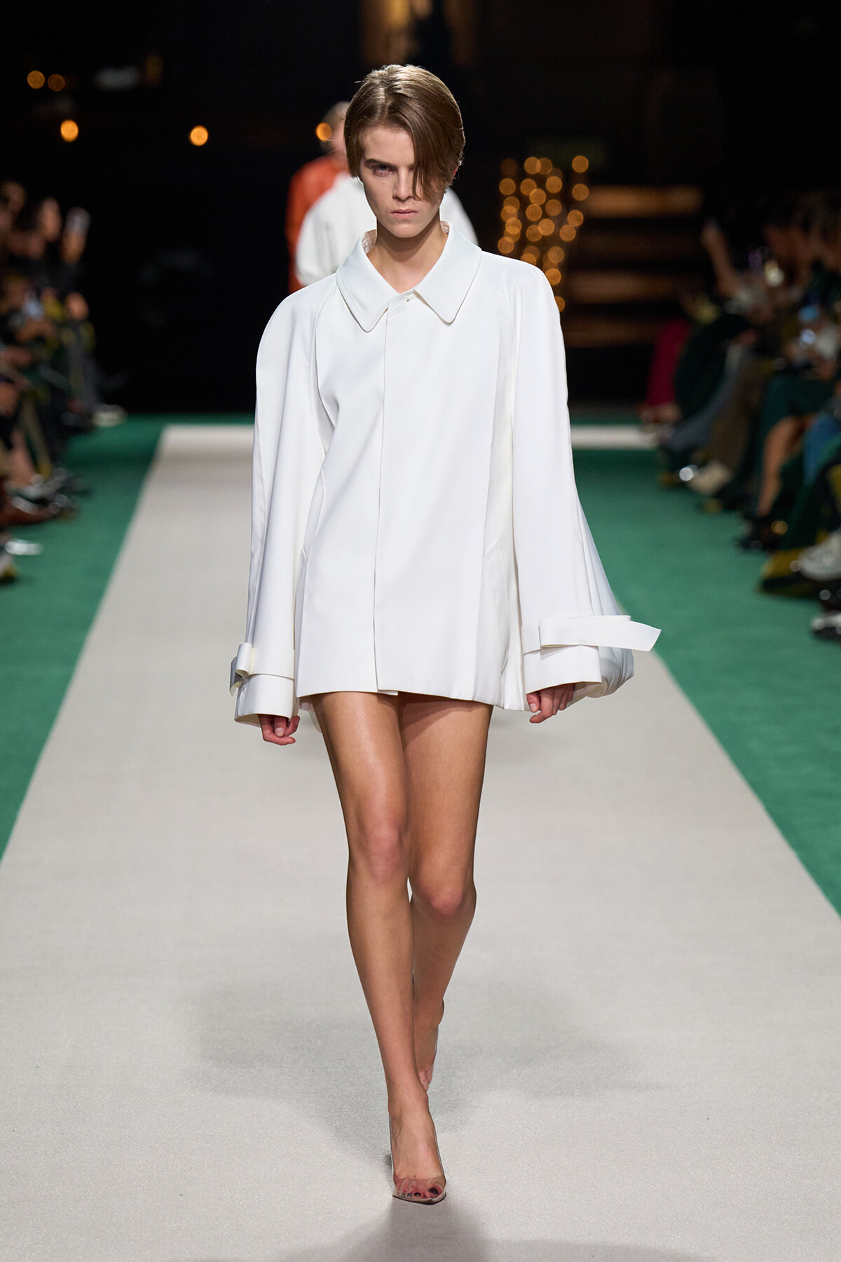 Model walking a fashion runway in an oversized white coat with wide cuffs and short hem, neutral backdrop.