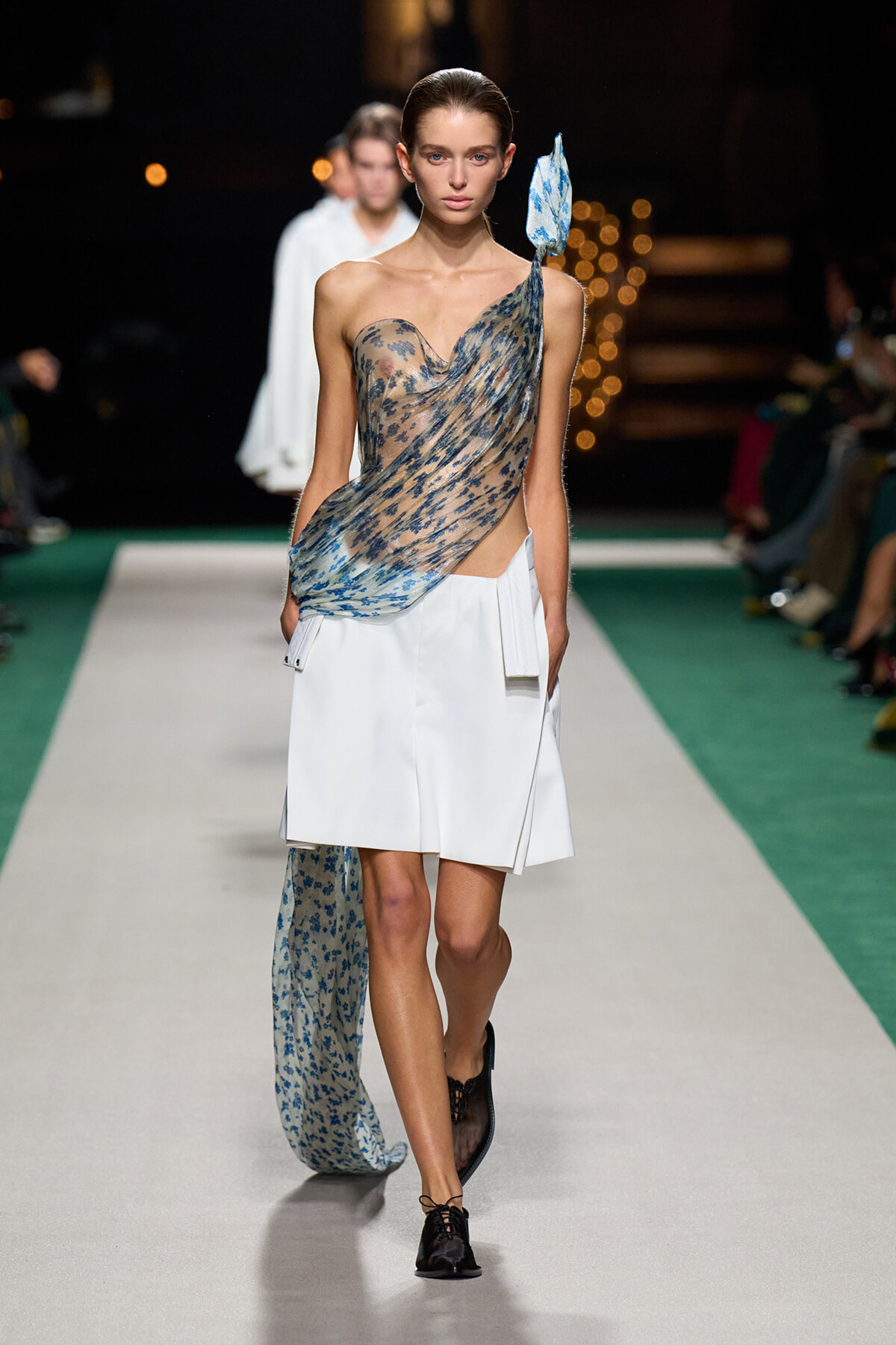 Model on a runway wearing a sheer blue floral draped one-shoulder top paired with a white structured skirt and a long trailing scarf fabric