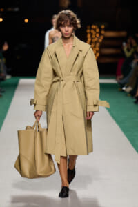 Model walks the runway in a tan oversized trench coat with a waist belt, carrying a large beige tote bag.