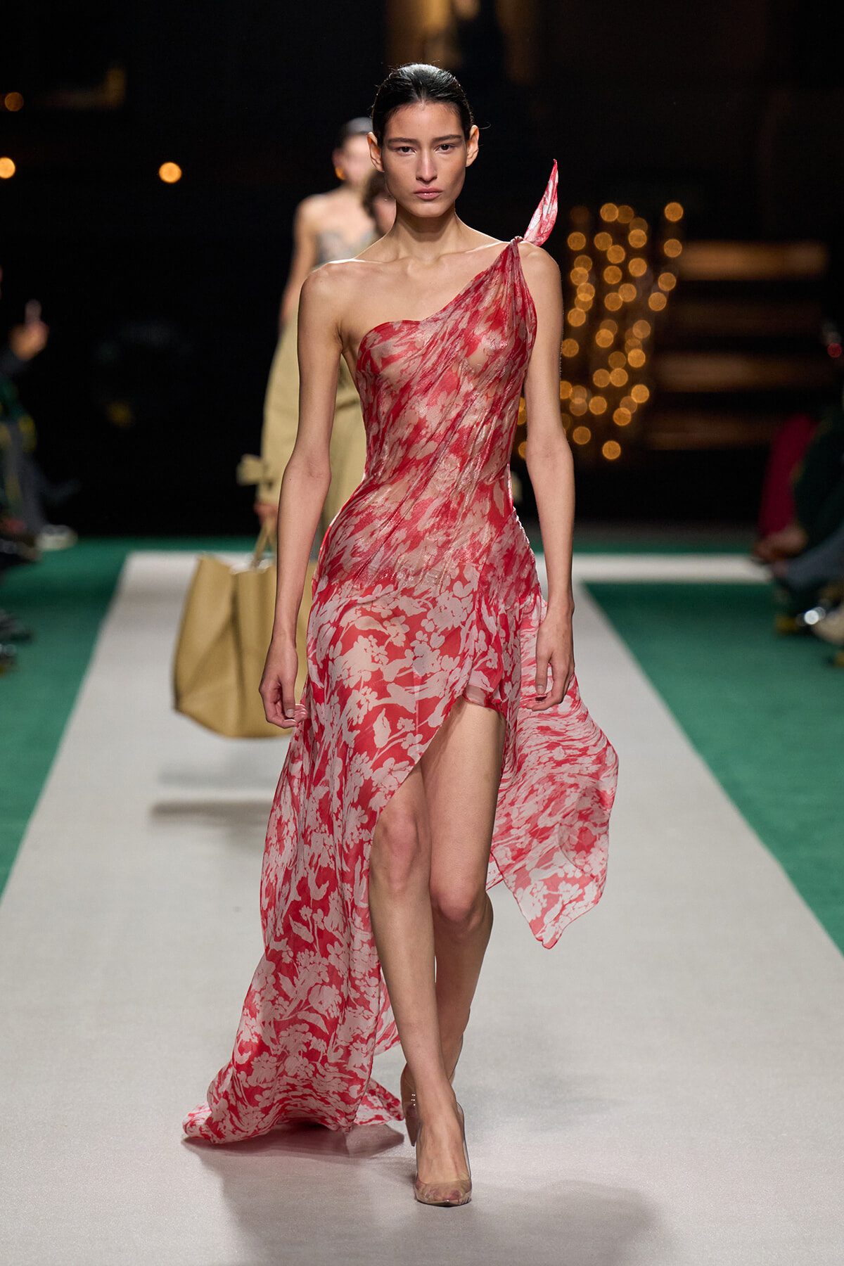 Model walking the runway in a pink-red floral one-shoulder gown with a high slit, under dark venue lights.