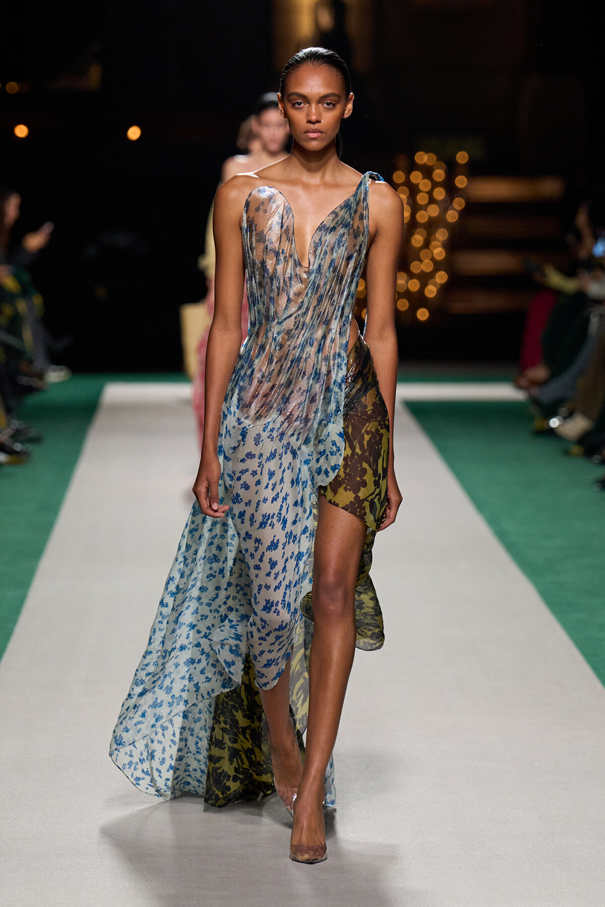 Model walks the runway in a sheer, blue floral gown with a corset-like bodice and layered, high-low skirt.