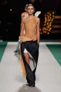 Runway model in a sheer peach draped one-shoulder top with black wide-leg pants and white shorts peeking out at the leg edge.
