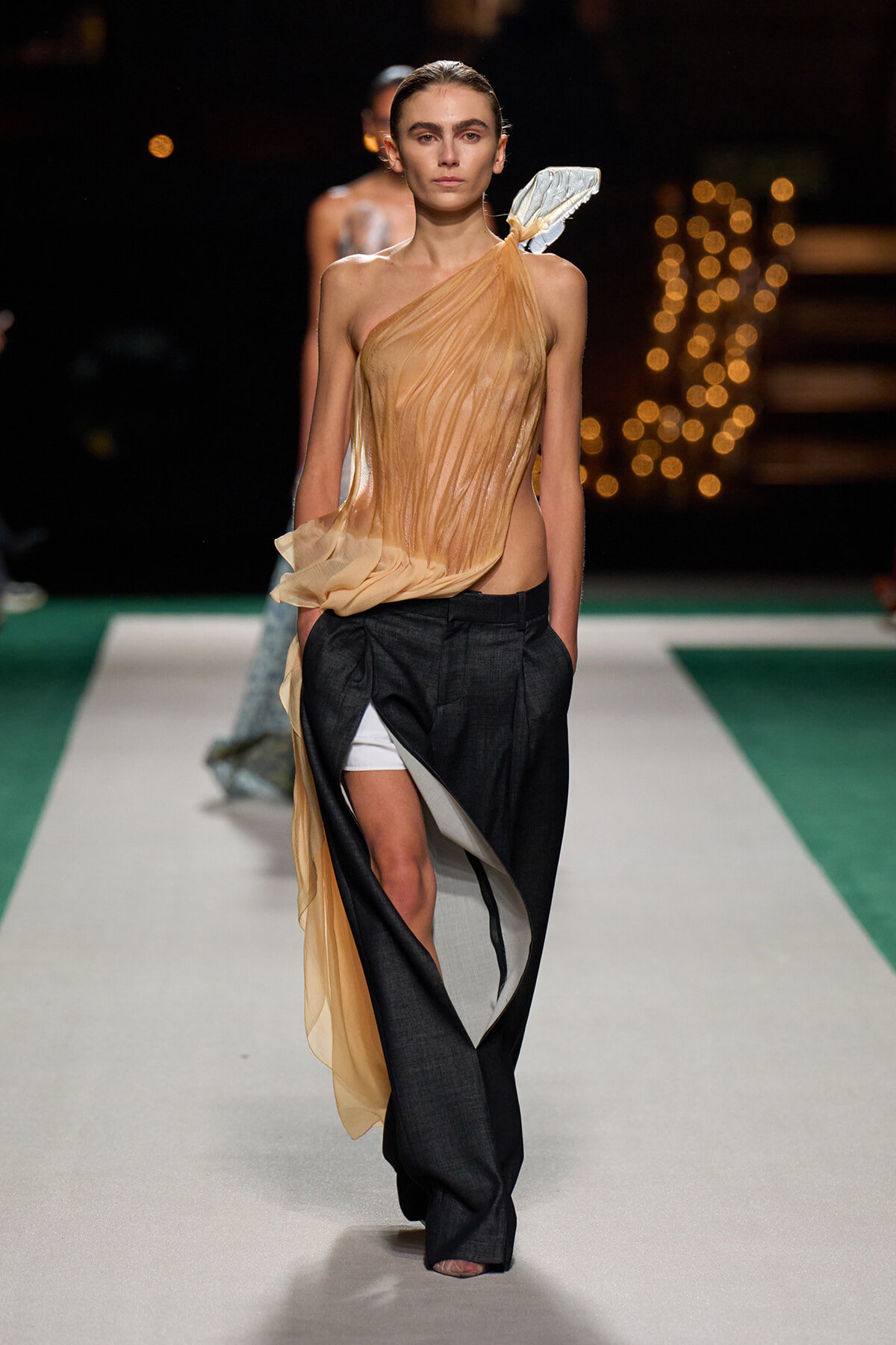 Runway model in a sheer peach draped one-shoulder top with black wide-leg pants and white shorts peeking out at the leg edge.