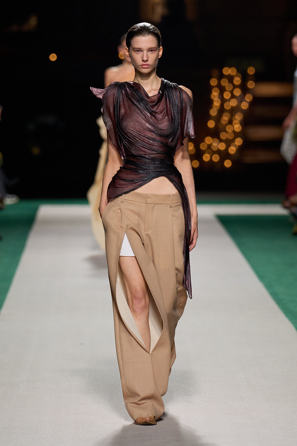 Model walking a runway in a sheer burgundy draped top and high-waisted tan wide-leg pants with inner white shorts showing through a side slit.