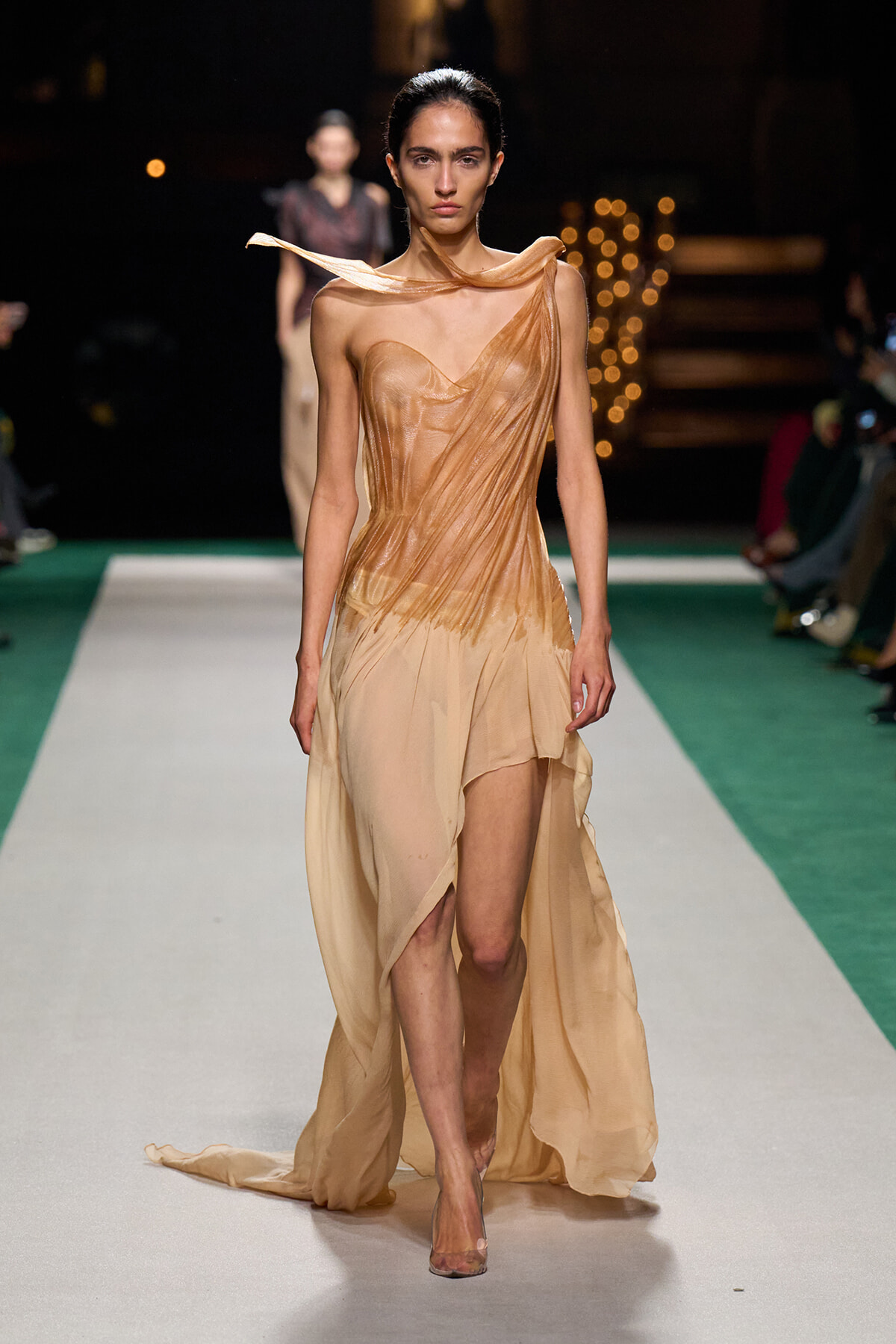 Model walks the runway in a tan, draped, one-shoulder gown with flowing train and sheer fabric details.