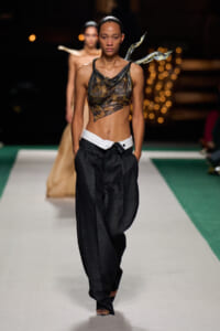 Model walking runway in a camo-draped sheer top and oversized dark trousers with a white belt.