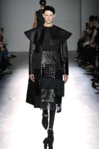 Model walks the runway in a black, patchwork ensemble featuring a dramatic oversized cape collar and mixed-texture panels.