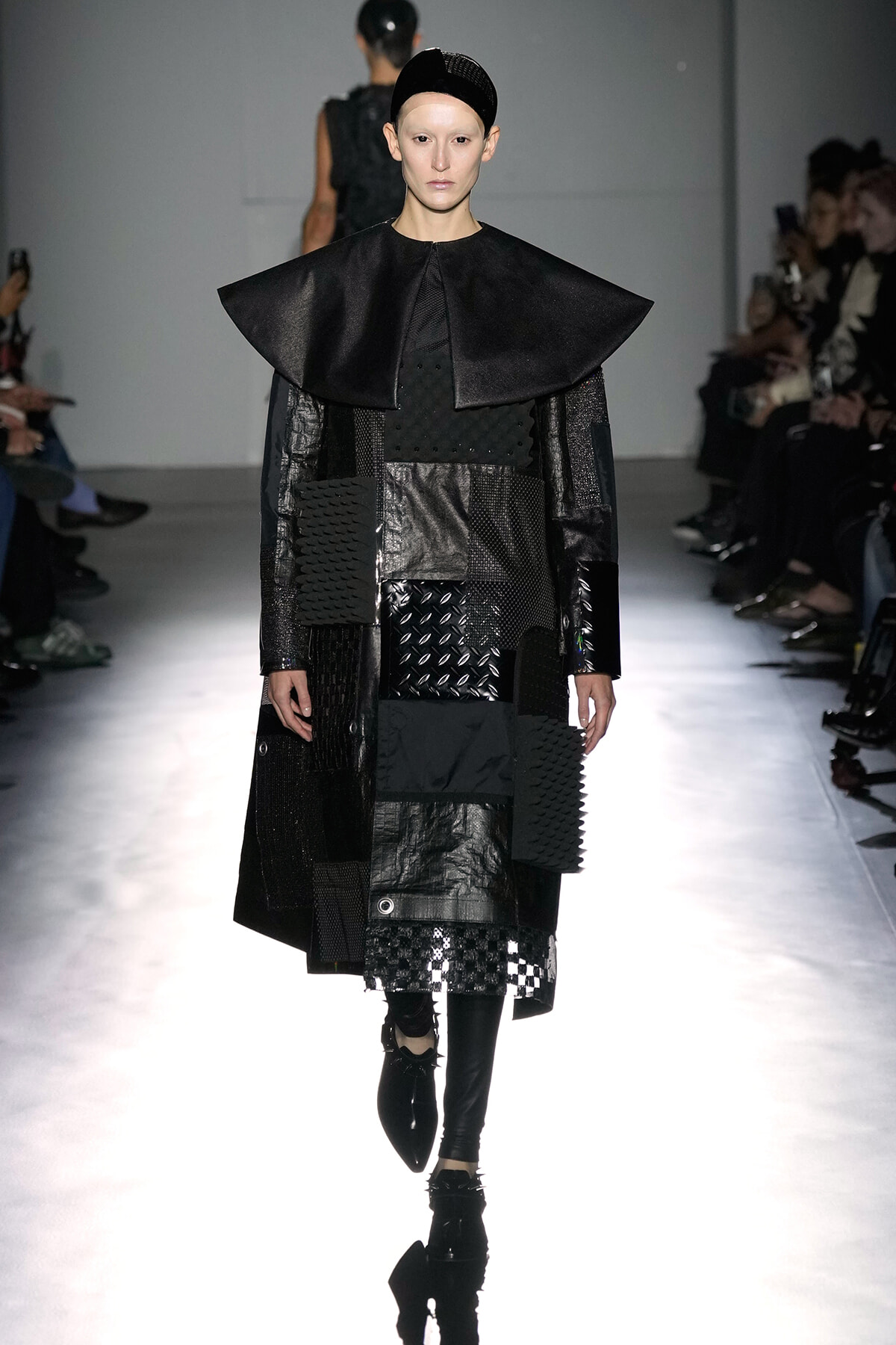 Model walks the runway in a black, patchwork ensemble featuring a dramatic oversized cape collar and mixed-texture panels.