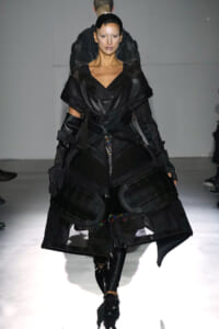 Model on a runway wearing a black, futuristic armor-inspired outfit with mesh panels and large, protective knee/hip guards.