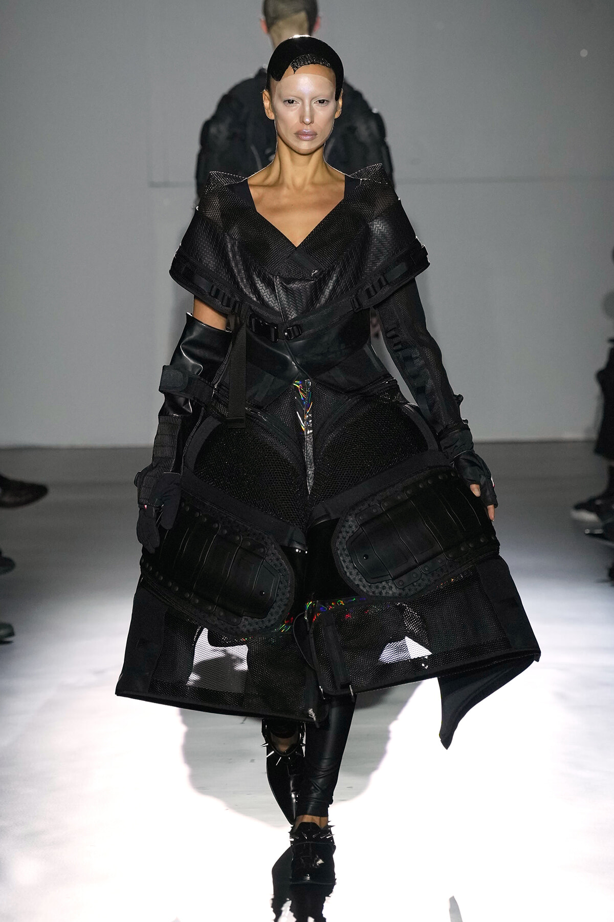 Model on a runway wearing a black, futuristic armor-inspired outfit with mesh panels and large, protective knee/hip guards.