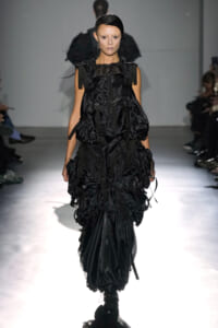 Female model walks the runway in an all-black, voluminous, strap-accented dress with gathered layers and utility details.