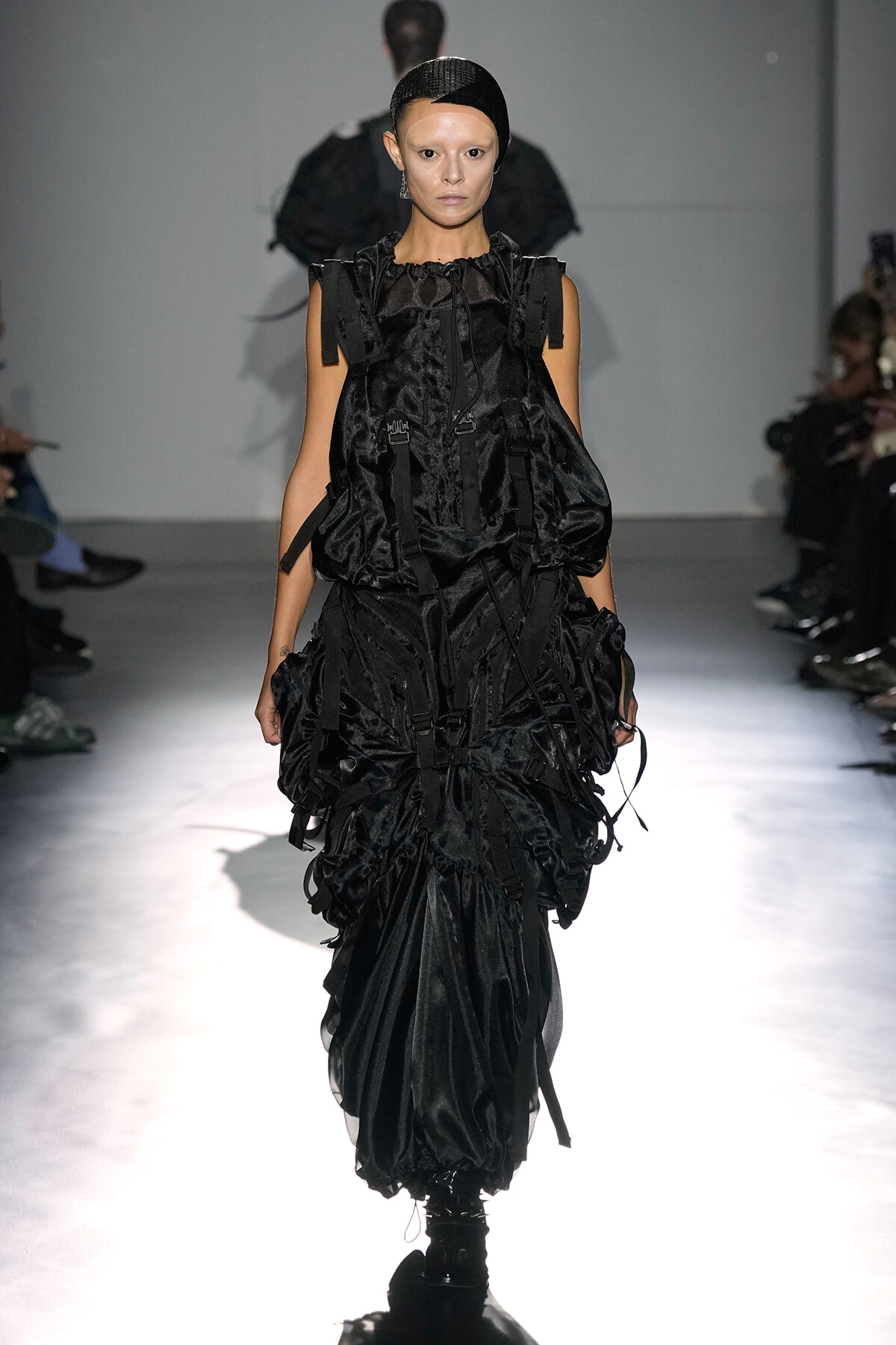 Female model walks the runway in an all-black, voluminous, strap-accented dress with gathered layers and utility details.