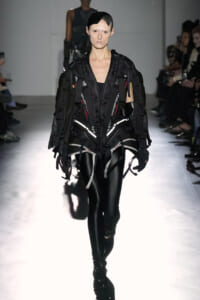 Runway model wearing an avant-garde black harness jumpsuit with straps and reflective accents, paired with black leggings and a mesh headpiece.