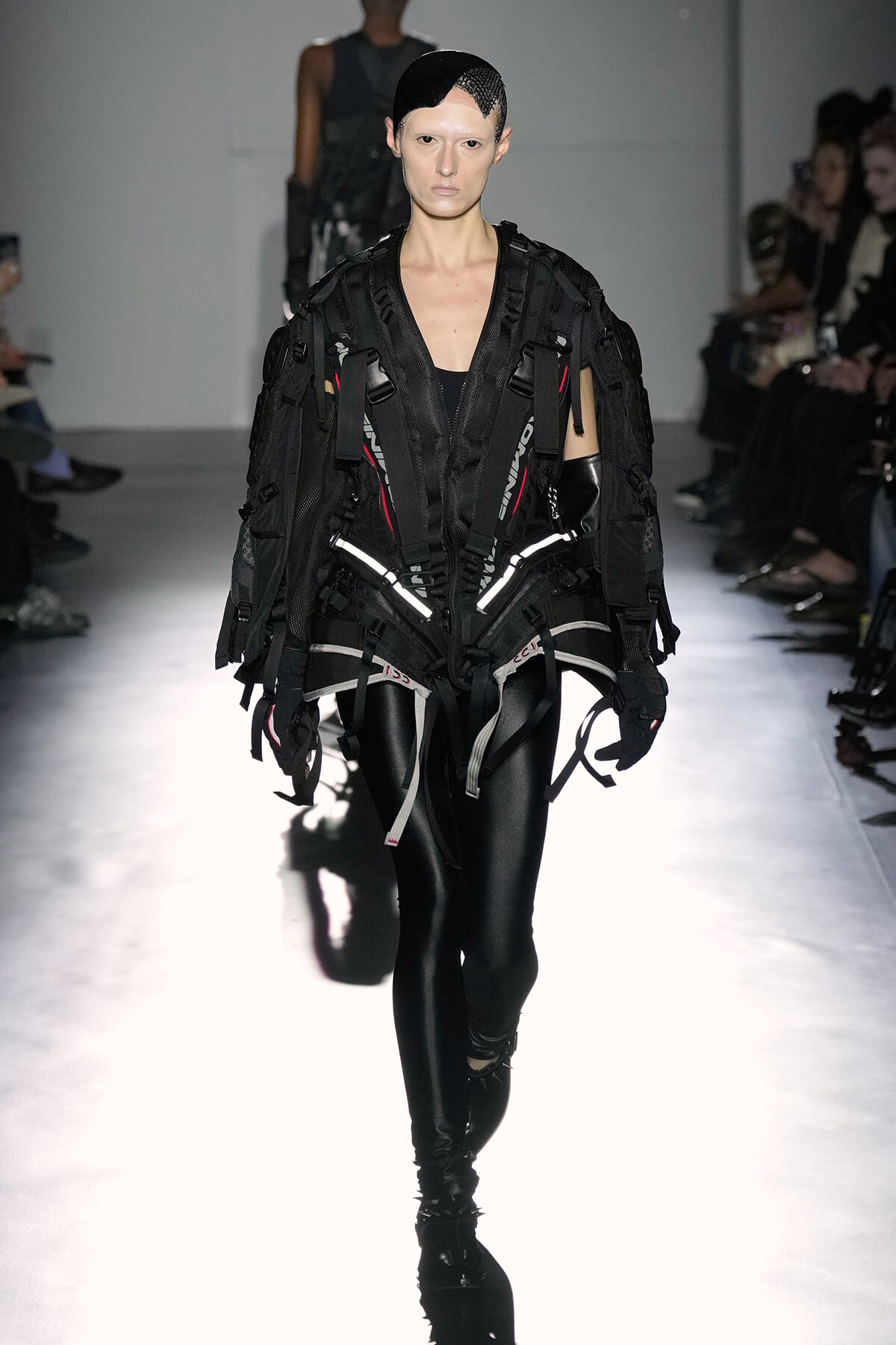 Runway model wearing an avant-garde black harness jumpsuit with straps and reflective accents, paired with black leggings and a mesh headpiece.