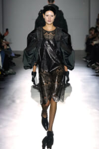 Model walks the runway in a black lace dress with sheer panels and dramatic oversized puff sleeves, paired with gloves and a glossy headband.