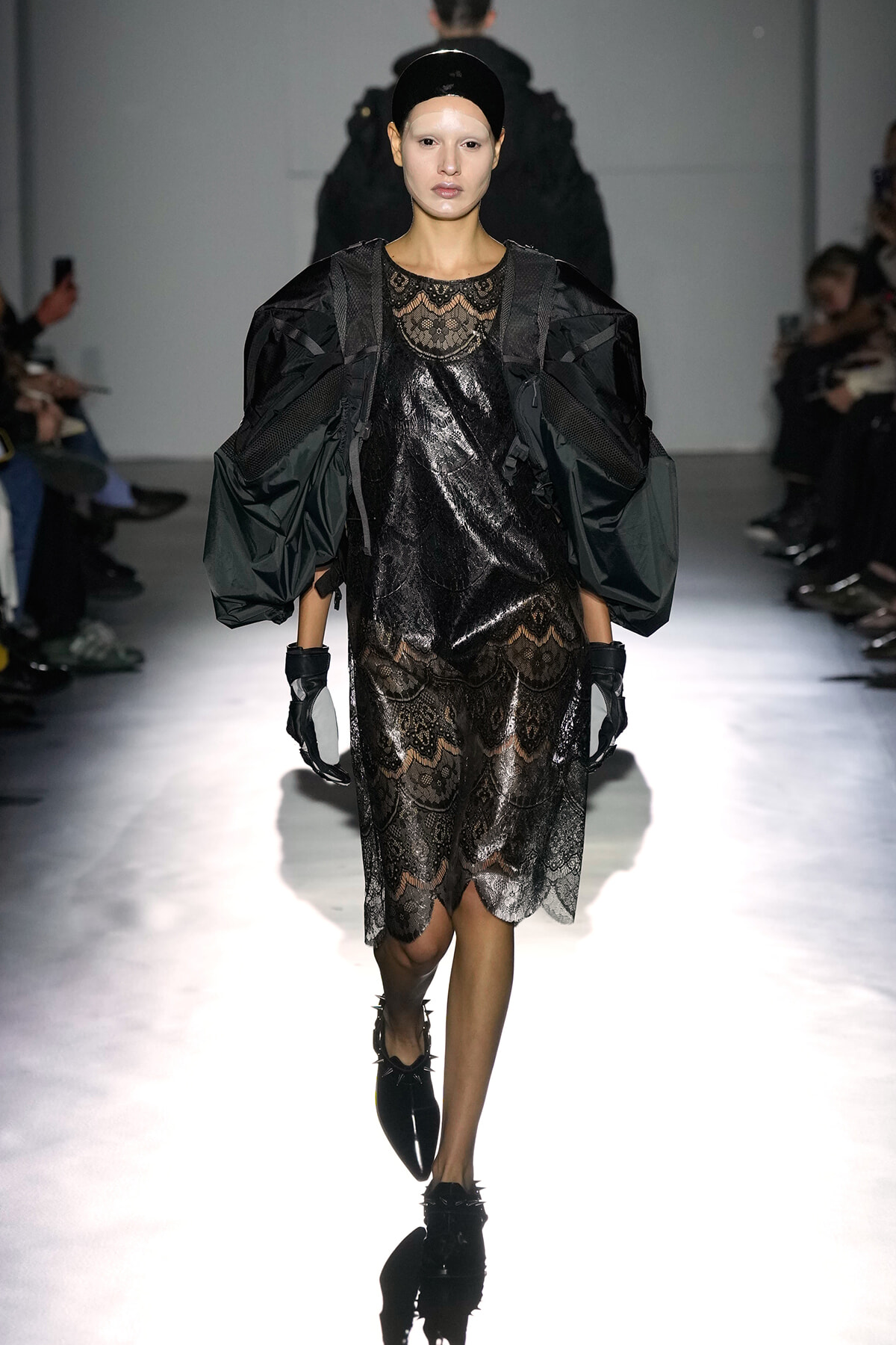 Model walks the runway in a black lace dress with sheer panels and dramatic oversized puff sleeves, paired with gloves and a glossy headband.