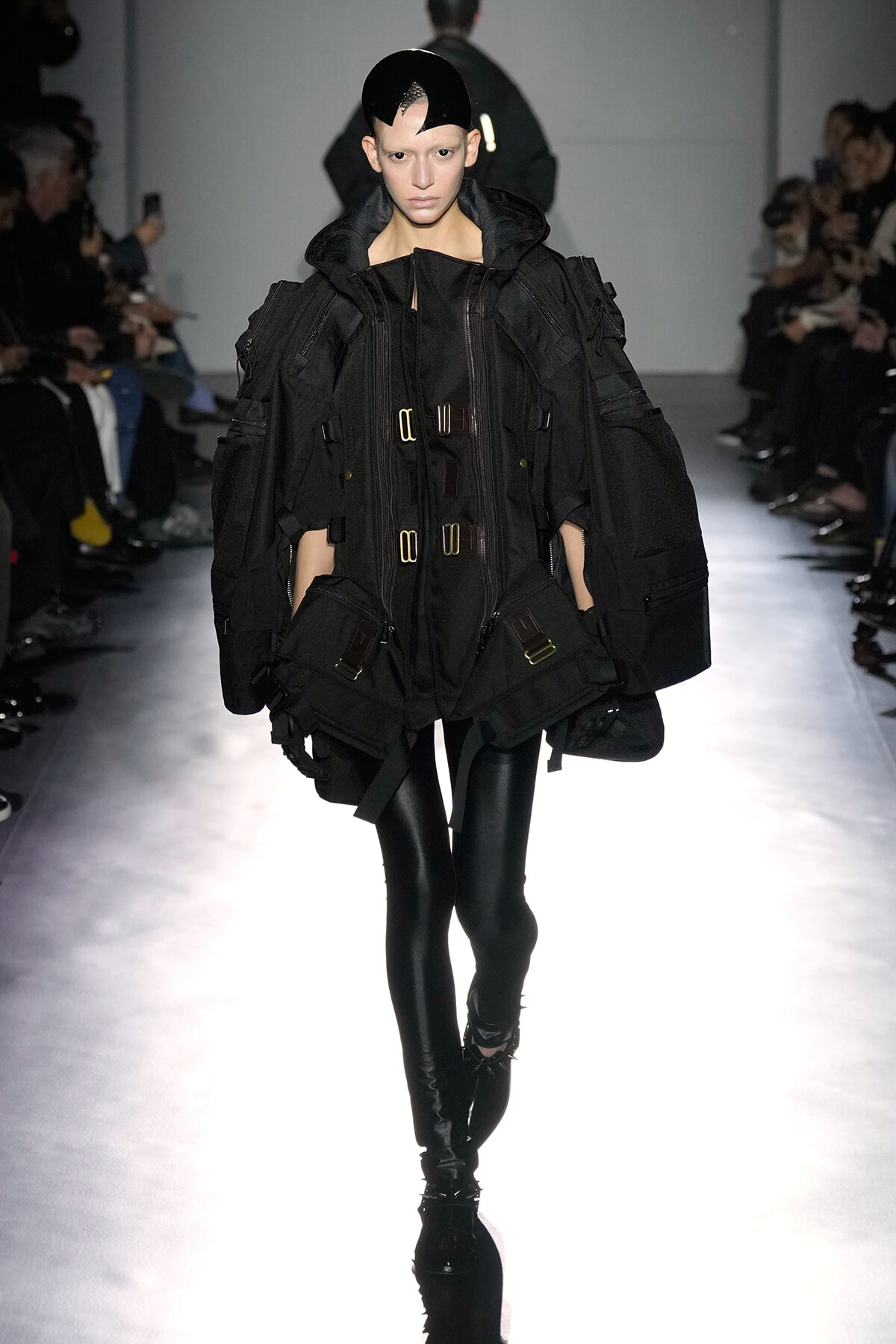 Model on a runway wearing an oversized black utility jacket with buckles and multiple pockets, paired with black leggings and boots.